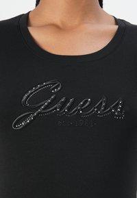 Black short-sleeve T-shirt featuring a glossy embroidered "Guess" logo embellished with metallic studs and "est. 1981" underneath.
