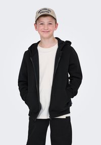 Sort zip-up hættetrøje med en stor hætte, båret over en lyse farvet t-shirt, parret med sorte bukser. En beige kasket med teksten "New York City Athletics".