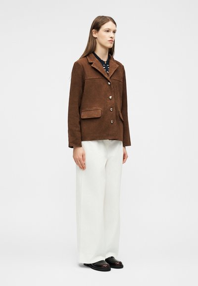 Γυναίκα που στέκεται φορώντας καφέ σακάκι με κουμπιά, μαύρο πουκάμισο, άσπρο παντελόνι με φαρδιά πόδια και μαύρα παπούτσια μπροστά σε απλό λευκό φόντο.