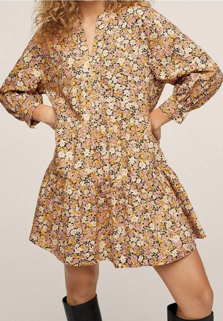 Femme portant une robe à manches longues imprimée de fleurs jaunes, roses et blanches, associée à des bottes noires. Mains sur les hanches.