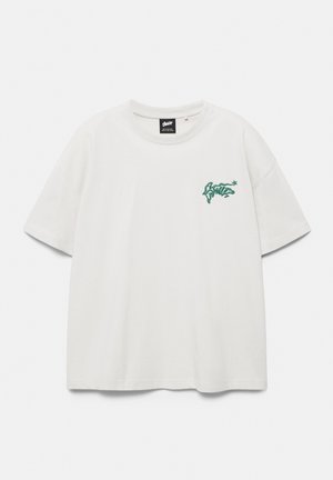 T-shirt blanc à manches courtes avec col rond, petit texte vert style graffiti et logo étoile sur la poitrine gauche.
