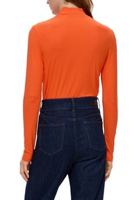 Osoba s priliehavým oranžovým topom s dlhým rukávom, zasunutým v nohaviciach z tmavomodrého denimu s vysokým pásom, stojí chrbtom k fotoaparátu.