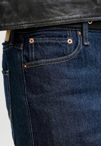 Mørkeblå denimjeanslomme med kobbernagler og kontrastsøm, kombinert med nederkant på en svart skinnjakke.