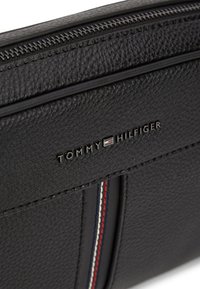 Μαύρο δερμάτινο πορτοφόλι με υφή, κλείσιμο με φερμουάρ και λογότυπο. Λεπτομέρειες περιλαμβάνουν κόκκινες, λευκές και μπλε αποχρώσεις κατά μήκος της πλευράς.