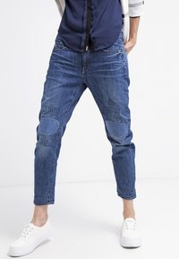 Vaqueros de denim en un color azul desgastado, con parches en las rodillas de un tono más oscuro y patrones sutiles. Estilo de corte holgado y con dobladillo remangado.