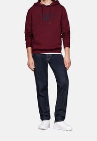 Mörkröd hoodie med marinblå logotyp, känguruficka och snörning; kombinerad med mörkblå jeans och vita sneakers. Enkelt, funktionellt design.