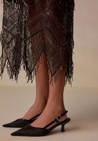 Black lace skirt with intricate geometric patterns and bead accents; paired with pointed-toe black slingback heels featuring a kitten heel.