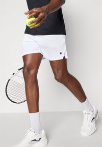 Shorts de sport blancs, accent logo noir, associés à des baskets blanches et des chaussettes à mi-mollet. Tient une balle de tennis verte, avec une raquette visible.