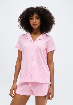 Žena s kučeravými vlasmi, ktorá má na sebe pyžamo so krátkym rukávom a gombíkmi v ružovo-bielych pásikoch a zodpovedajúce šortky, stojaca pred jednoduchým pozadím.