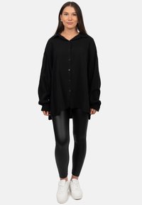 Camicia nera oversize con bottoni, realizzata in un tessuto strutturato e con colletto, abbinata a leggings neri lucidi e sneaker bianche.