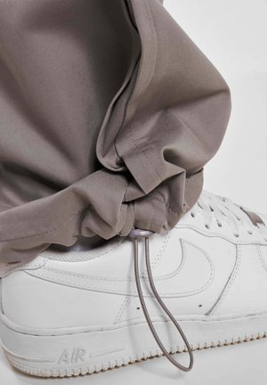 Világosszürke jogger nadrág, húzózsinórral az alján, párosítva fehér sportcipőkkel, amelyek textúrázott talppal és jellegzetes logó részlettel rendelkeznek.