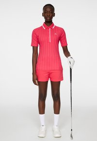 Różowa koszulka polo z krótkimi rękawami, w teksturowane paski i z białym kołnierzykiem; w zestawie z pasującymi różowymi szortami. Trzyma kij golfowy.