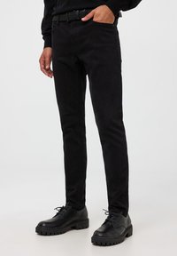 Jeans neri slim-fit in denim, con una texture liscia, tasche standard e abbinati a scarpe nere con lacci su uno sfondo neutro.