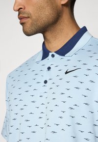 Light blue polo shirt with a navy collar, featuring a black Nike logo and small dark patterns throughout. Short sleeves and button placket.