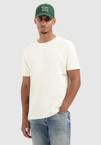 Crème korte mouwen t-shirt met een ontspannen pasvorm, voorzien van een zwart gedrukt logo aan de linkerzijde. Gepaard met lichtblauwe denim jeans.