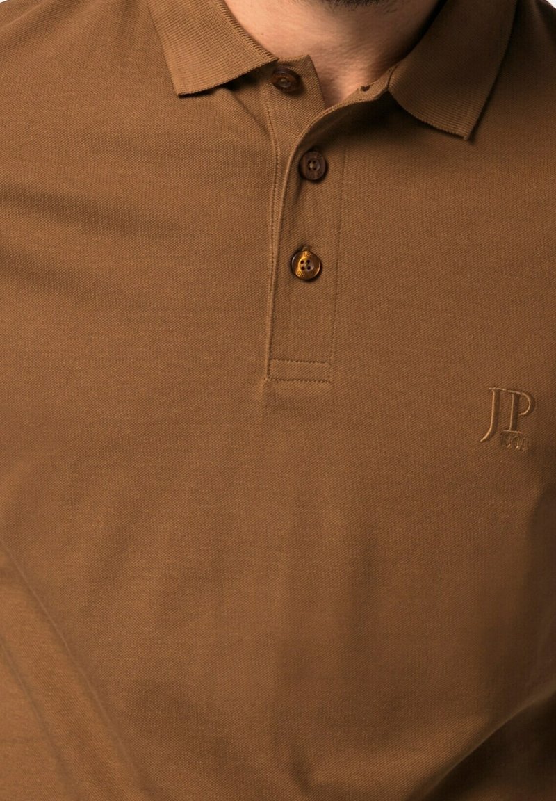 Polo marron avec trois boutons et petit logo brodé "JP 1931" sur la poitrine gauche, porté par une personne.