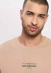 Beżowa bawełniana koszulka z krótkim rękawem, z nadrukowanym hasłem w kolorze czarnym: "NIGDY NIE JEST ZA PÓŹNO NA START" z logo "CROSS JEANS" poniżej.