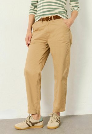 Person trägt beige Chinohose, braunen Gürtel, grün-weiß gestreiftes Langarmshirt und beige Sneaker, steht mit einer Hand in der Tasche.