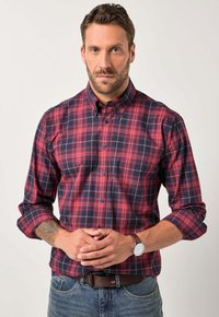 Rood en marineblauw geruit overhemd met knopen en lange mouwen, voorzien van een button-down kraag en contrasterende knopen. Draagt een bruine riem en horloge.