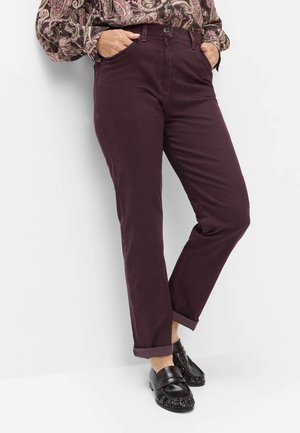 Person iført optrukne mørkelilla bukser, sorte loafers og en mønstret bluse med lange ærmer med hænderne i lommerne.