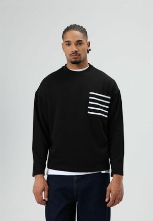 Zwarte trui met lange mouwen en een wit gestreept borstzakje. Heeft een losse pasvorm en een ronde hals, gecombineerd met blauwe spijkerbroek.