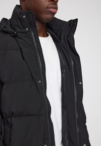 Schwarze gepolsterte Jacke mit hohem Kragen, Reißverschluss und Seitentaschen. Der Stoff wirkt glatt mit matter Oberfläche.