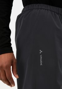 Pantalons imperméables noirs avec une taille élastique et le logo "VAUDE" en gris clair sur le côté. Texture de tissu lisse et durable.