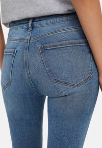 Jeans de mezclilla azul con un diseño ajustado, que cuentan con bolsillos traseros estándar y detalles de costura visibles. La tela muestra un ligero desvanecimiento y textura.