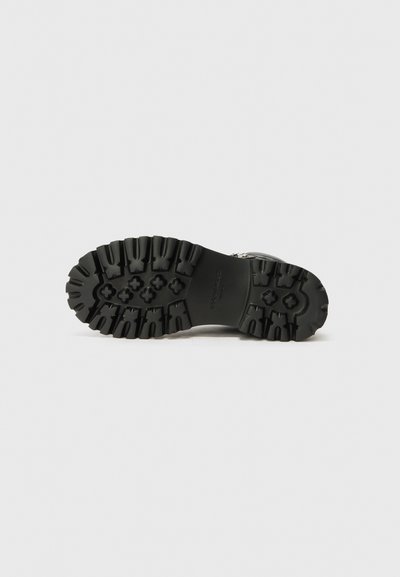 Suela de zapato de goma negra con surcos profundos. Superficie texturizada y ranuras pronunciadas para mejorar la tracción. Presenta una marca grabada en la zona del talón.