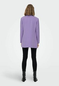 Violetti pitkä bleiseri, jossa on muotoiltu leikkaus, yksittäinen takakara ja pitkät hihat. Päälle puettuna mustat legginssit ja nilkkurit, neutraalia taustaa vasten.