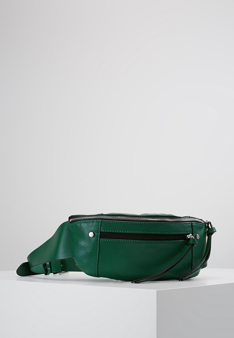 Banane en cuir vert foncé avec fermetures éclair argentées, poche avant et sangle réglable reposant sur une surface blanche face à un fond gris.