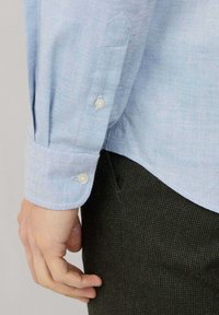 Camicia a rombi di colore blu chiaro con colletto a punta, polsini con bottone e bottoni a contrasto, abbinata a pantaloni verde scuro con texture.