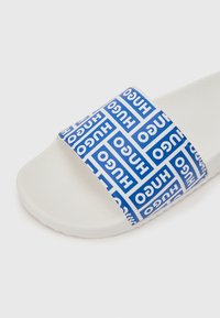 Λευκή σαγιονάρα με φαρδιά μπλε λουρί καλυμμένο με επαναλαμβανόμενα λευκά λογότυπα "HUGO" σε διάφορες κατευθύνσεις.