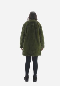 Cappotto in pelo finto verde oliva, maniche lunghe, colletto grande, vestibilità rilassata, lunghezza fino a metà coscia, indossato sopra leggings neri e scarpe chunky nere.