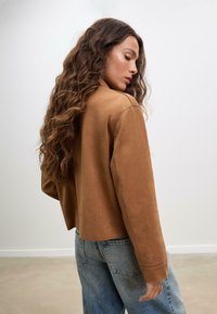 Veste cropped en daim marron avec un col et une texture unie, associée à un jean en denim taille haute bleu clair. Vue de dos avec des cheveux ondulés longs.