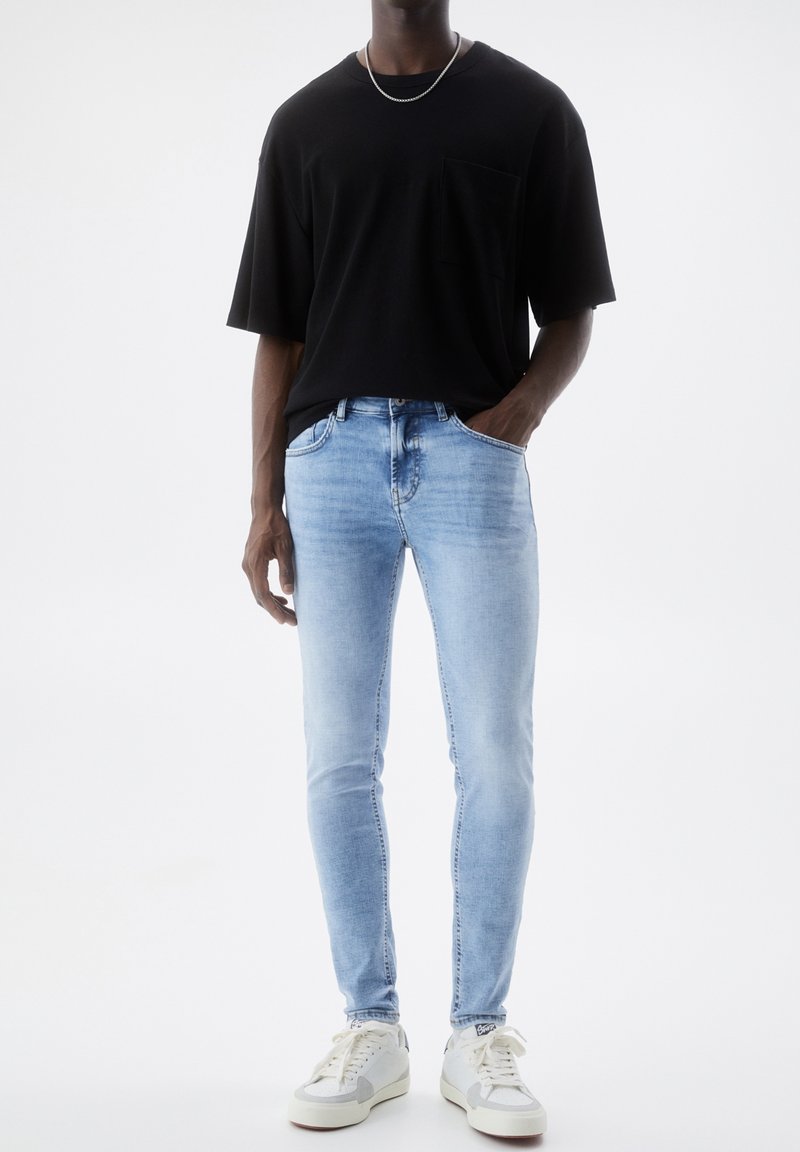Homme portant un jean slim bleu clair, un t-shirt noir oversize, des baskets blanches et une chaîne argentée, debout avec une main dans la poche.