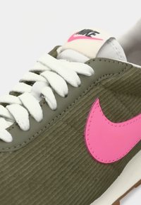 Zaļas kordūras sporta apavus ar baltām auklām, rozā Nike swoosh logotipu, teksturētu virsu un krēmīgu mēli. Iezīmēta šuvju apdare un gumijas zole.