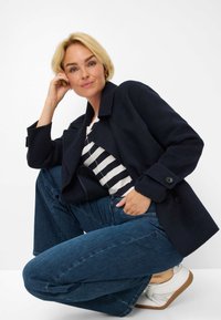 Blazer in misto lana di colore navy con bottoni grandi, top a strisce bianco e nero, jeans blu e sneakers bianche. Modello accovacciato, posando con sicurezza.