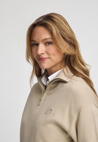 Beżowy sweter z półzamkiem i teksturowanym materiałem. Posiada małe, haftowane logo na klatce piersiowej oraz klasyczny krój.