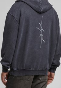 Donkergrijze hoodie met een ontspannen pasvorm. Heeft een verticale abstracte vormgeving in lichtgrijs op de achterkant. Zachte stofstructuur.