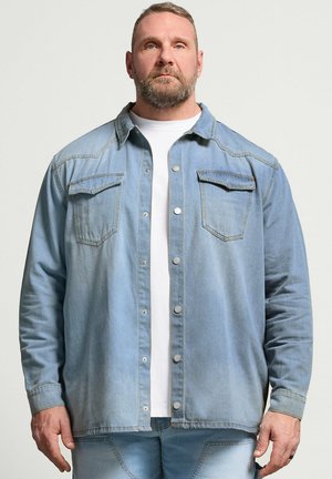 Mężczyzna z brodą, ubrany w jasnoniebieską kurtkę jeansową na białej koszuli, stojący na jednolitym jasnym tle, zwrócony przodem.