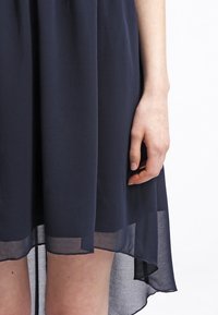 Robe bleu marine en tissu léger, avec un ourlet fluide et une finition transparente, avec une main tenant le tissu sur le côté.