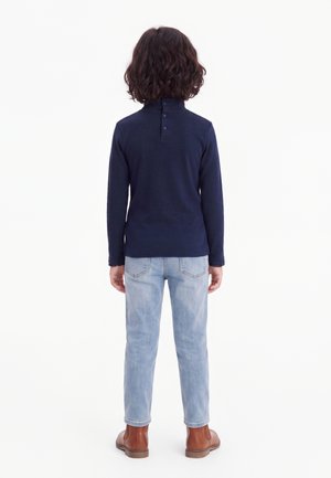 Child with curly dark hair wears a navy long-sleeve top, light blue jeans, and brown ankle boots, standing facing away on a white background.