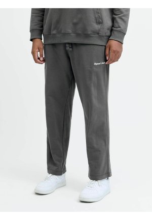 Person iført mørkegrå joggingbukser med lille hvid tekst på låret og hvide sneakers, stående mod en ensfarvet lys baggrund.