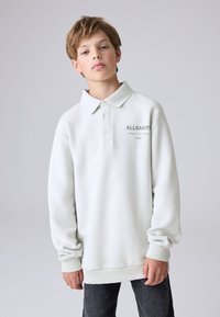 Teenager s světle hnědými vlasy, oblečen�ý v bílé dlouhém rukávu polotriku ALLSAINTS a tmavých džínách na jednobarevném šedém pozadí.