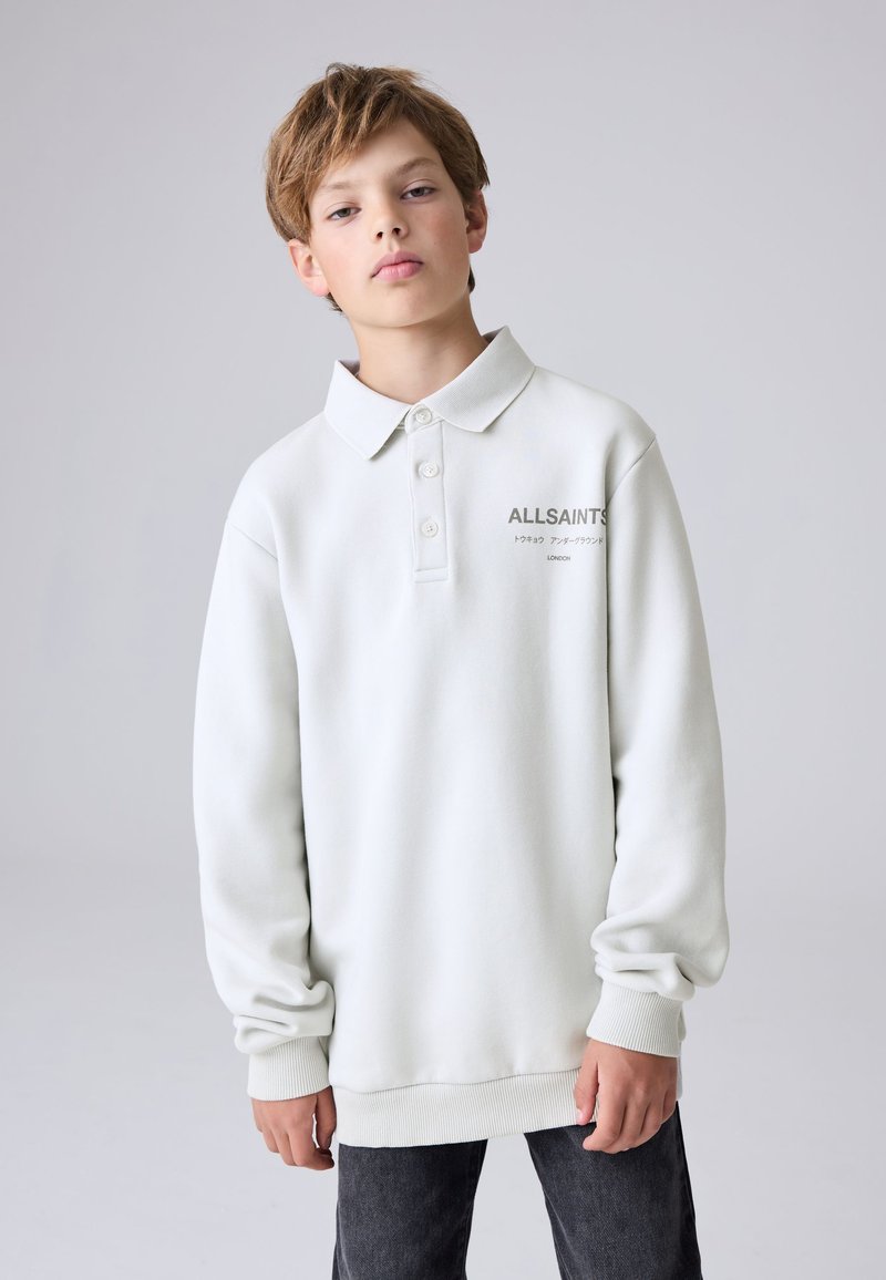 Teenager s světle hnědými vlasy, oblečený v bílé dlouhém rukávu polotriku ALLSAINTS a tmavých džínách na jednobarevném šedém pozadí.