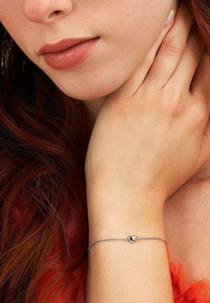 Close-up of a woman's wrist wearing a delicate silver chain bracelet with a small heart charm and her hand gently touching her neck.