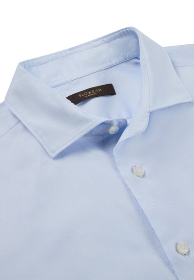 Camisa de color azul claro con cuello, hecha de tela suave, con textura lisa, tres botones y una etiqueta tejida con "SLOWEAR" sobre un fondo marrón.