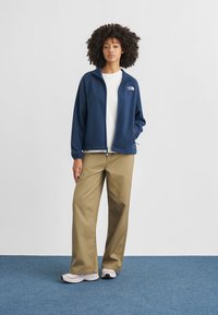 Νεαρή γυναίκα με σγουρά μαλλιά που φοράει μπλε στρουμπουλό μπουφάν με φερμουάρ, λευκό πουκάμισο, μπεζ παντελόνια φαρδιάς γραμμής και λευκά αθλητικά παπούτσια, στέκεται σε μπλε χαλί.