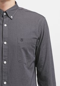 Camicia button-up grigia scura in cotone con taschino sul petto, bottoni bianchi e colletto. Presenta un logo sottile sulla tasca.